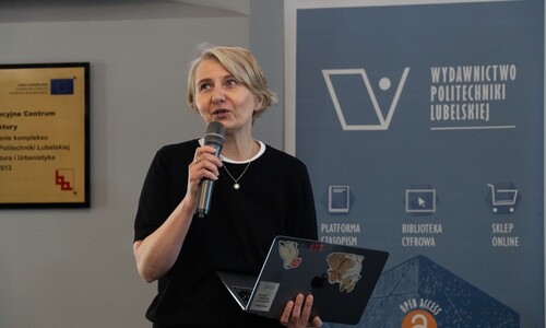 Kobieta o blond włosach przemawia do mikrofonu, trzymając w dłoniach otwartego laptopa z naklejkami. Obok niej znajduje się roll-up Wydawnictwa Politechniki Lubelskiej.