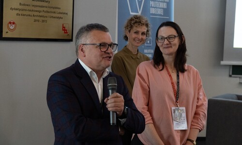 Mężczyzna w marynarce w kratę przemawia do mikrofonu. Obok niego, nieco z tyłu, uśmiechają się dwie kobiety.