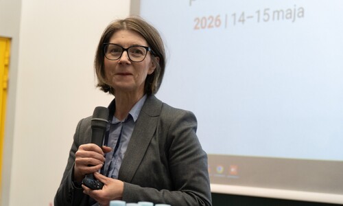 Kobieta w okularach i szarej marynarce przemawia do mikrofonu podczas konferencji, stojąc na tle ekranu wyświetlającego tekst.