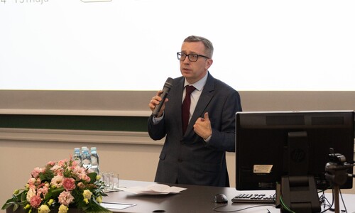 Mężczyzna w garniturze i krawacie przemawia do mikrofonu, gestykulując dłonią. Na stole przed nim znajduje się bukiet r&oacute;żowych kwiat&oacute;w oraz monitor.