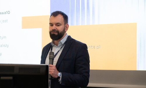 Mężczyzna z brodą w granatowej marynarce i jasnej koszuli przemawia do mikrofonu podczas prezentacji, stojąc przed ekranem z tekstem.