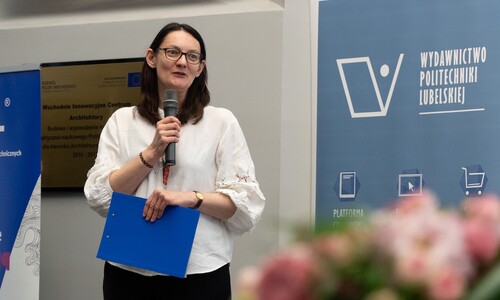 Kobieta w białej bluzce przemawia do mikrofonu, trzymając w ręku niebieską teczkę. Obok niej widoczny jest roll-up Wydawnictwa Politechniki Lubelskiej.