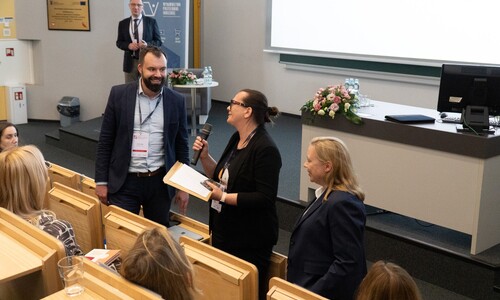 Widok na salę wykładową. Kobieta z mikrofonem rozmawia z mężczyzną stojącym w przejściu między rzędami foteli. Obok nich stoi trzecia kobieta, a w tle przy stole widać siedzących uczestnik&oacute;w.