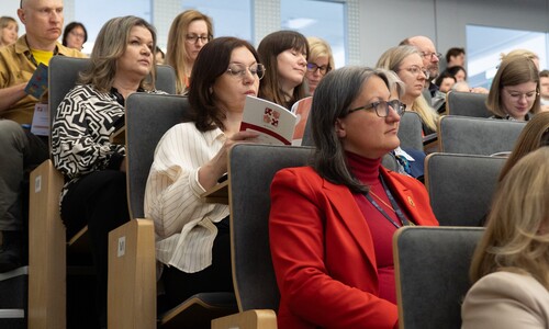 Uczestnicy konferencji siedzący w sali wykładowej na fotelach. W centrum kadru kobieta w czerwonej marynarce uważnie przysłuchuje się wystąpieniu, obok niej inna uczestniczka przegląda materiały konferencyjne.