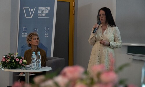 Kobieta w jasnej sukience przemawia do mikrofonu, stojąc obok stolika, przy kt&oacute;rym siedzi moderatorka panelu. W tle widoczny jest roll-up Wydawnictwa Politechniki Lubelskiej.