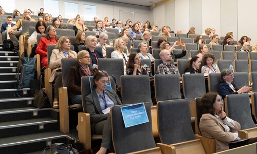 Widok na wypełnioną salę wykładową podczas konferencji. Uczestnicy siedzą w rzędach foteli, niekt&oacute;rzy robią zdjęcia lub przeglądają materiały.