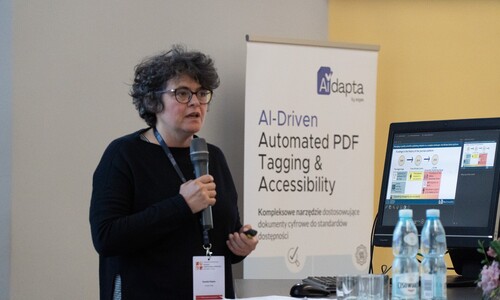 Kobieta w ciemnym swetrze i okularach przemawia do mikrofonu podczas prezentacji. Obok niej znajduje się roll-up z napisem &bdquo;AI-Driven Automated PDF Tagging & Accessibility&rdquo; oraz monitor z wyświetlonymi wykresami.