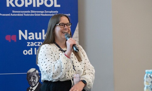 Kobieta w okularach i biało-czarnej bluzce w kropki uśmiecha się, przemawiając do mikrofonu. W tle widoczny jest niebieski roll-up stowarzyszenia Kopipol.