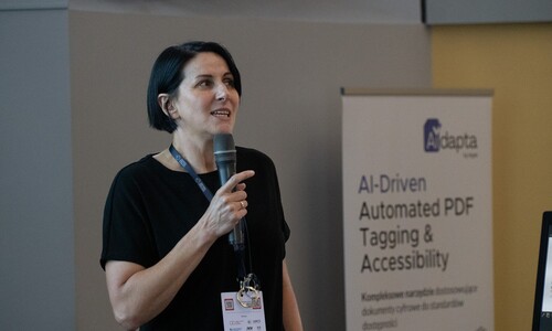 Kobieta z kr&oacute;tkimi ciemnymi włosami w czarnej bluzce przemawia do mikrofonu. Obok niej znajduje się roll-up z napisem &bdquo;AI-Driven Automated PDF Tagging & Accessibility&rdquo;.