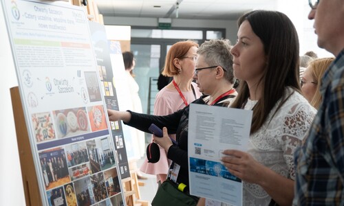 Uczestnicy konferencji przeglądają plakat naukowy zatytułowany &bdquo;Otwarty Szczecin &ndash; Cztery uczelnie, wsp&oacute;lne dane, nowe możliwości&rdquo;. Kobieta w białej bluzce wskazuje palcem na treść plakatu.