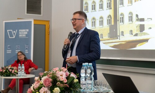 Mężczyzna w ciemnej marynarce przemawia do mikrofonu podczas prezentacji. Obok przy stole siedzi kobieta w czerwonym stroju, a w tle widać ekran z fotografią budynku.