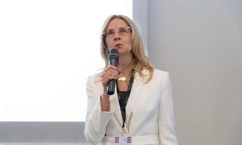 Kobieta w eleganckim, jasnym kostiumie przemawia do mikrofonu podczas wystąpienia konferencyjnego.