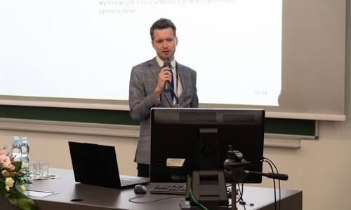 Mężczyzna w niebieskiej marynarce w kratę przemawia do mikrofonu, stojąc za stołem z laptopem i monitorem.